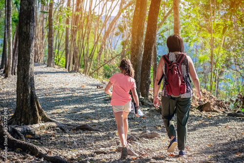 Back view of young girl and her mother walking together in forest nature path walk on trail woods background at Hellfire Pass in Kanchanaburi Thailand. Happy people relaxing on active outdoor activity photo