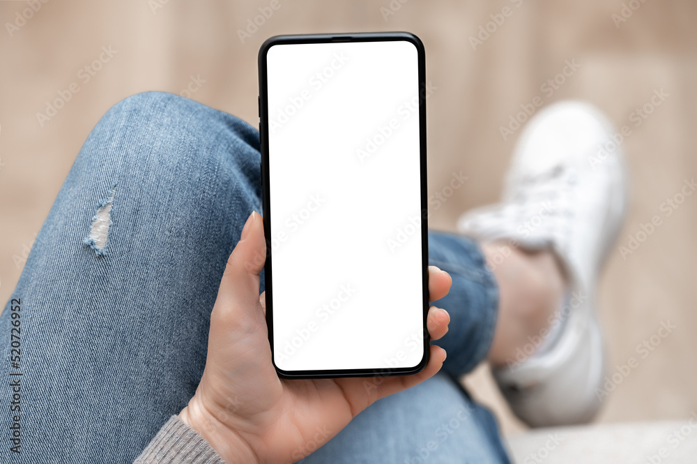 Top view mockup image of woman holding mobile phone with blank white screen. Woman in jeans sits cross-legged and holds cell phone in her hands with blank white screen
