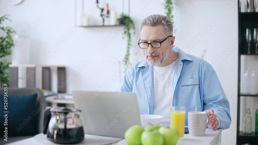 Busy man in his 50s working on laptop while having breakfast, multi-tasking Busy man in his 50s working on laptop while having breakfast, multi-tasking