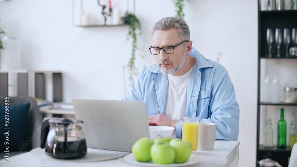 Middle-aged man having breakfast and reading news on laptop, morning routine Middle-aged man having breakfast and reading news on laptop, morning routine