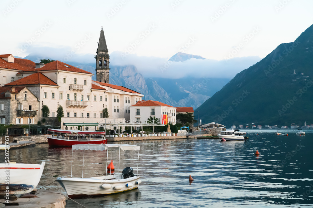 View of the bay in Perast. Red roofs, boats, tower, restaurant, sunset, mountains in the clouds in the background.