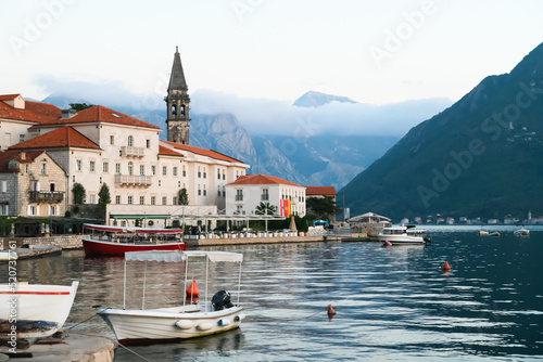 View of the bay in Perast. Red roofs, boats, tower, restaurant, sunset, mountains in the clouds in the background. © AnnFossa