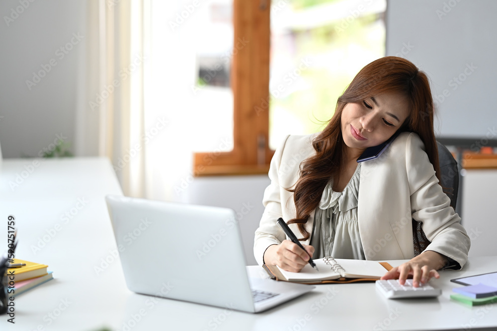 Busy millennial working woman negotiating with client via mobile phone and making notes on notebook Busy millennial working woman negotiating with client via mobile phone and making notes on notebook