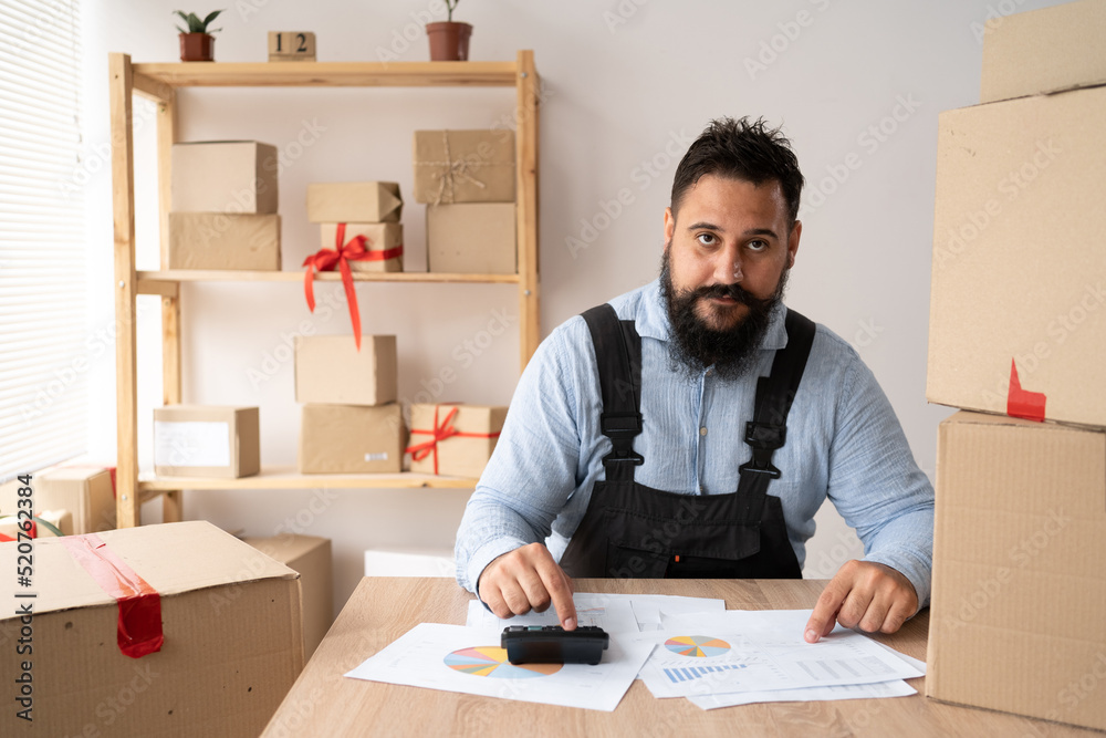 Portrait of Indian man, e-commerce employee sitting in the office full of boxes on the table using a calculator, SME, e-commerce, technology and delivery business. Portrait of Indian man, e-commerce employee sitting in the office full of boxes on the table using a calculator, SME, e-commerce, technology and delivery business.