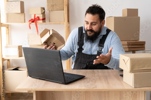 Evil bearded male business owner sitting at table and calling while browsing on laptop. sme business freelance working in home office using computer, online marketing packaging box delivery,