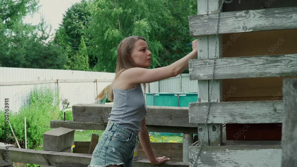 A beautiful caucasian girl in a gray tank top and short denim shorts paints the house with a brush. A European woman paints an old porch with white paint. painting work in a country house. A beautiful caucasian girl in a gray tank top and short denim shorts paints the house with a brush. A European woman paints an old porch with white paint. painting work in a country house.