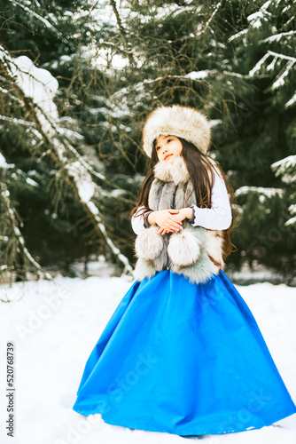 portrait of beautiful cute little Asian girl in fur coat and fur hat standing in snowy forest in winter, winter holidays and having fun, Christmas vacation © klavdiyav