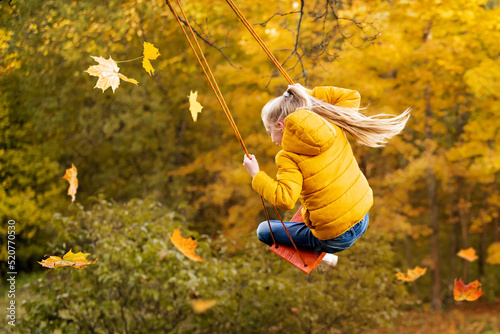 Happy little blonde caucasian girl smiling and riding a rope swing in autumn in the park.