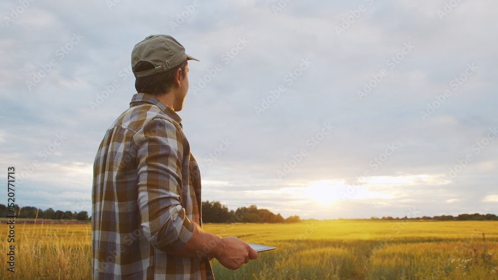 Farmer with a tablet computer in front of a sunset agricultural landscape. Man in a countryside field. Country life, food production, farming and technology.