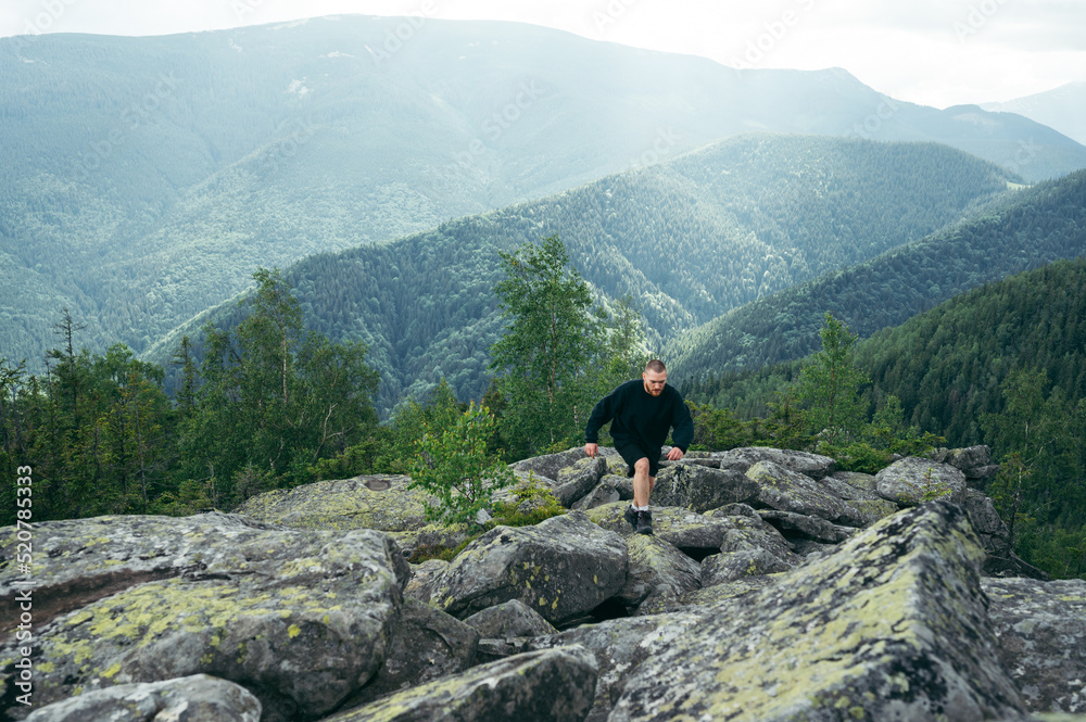 Focus on the landscape. Active young man in casual clothes is climbing a rock in the mountains at the top and looking at the camera.