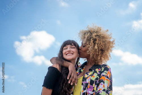 Woman giving girlfriend kiss on cheek against blue sky