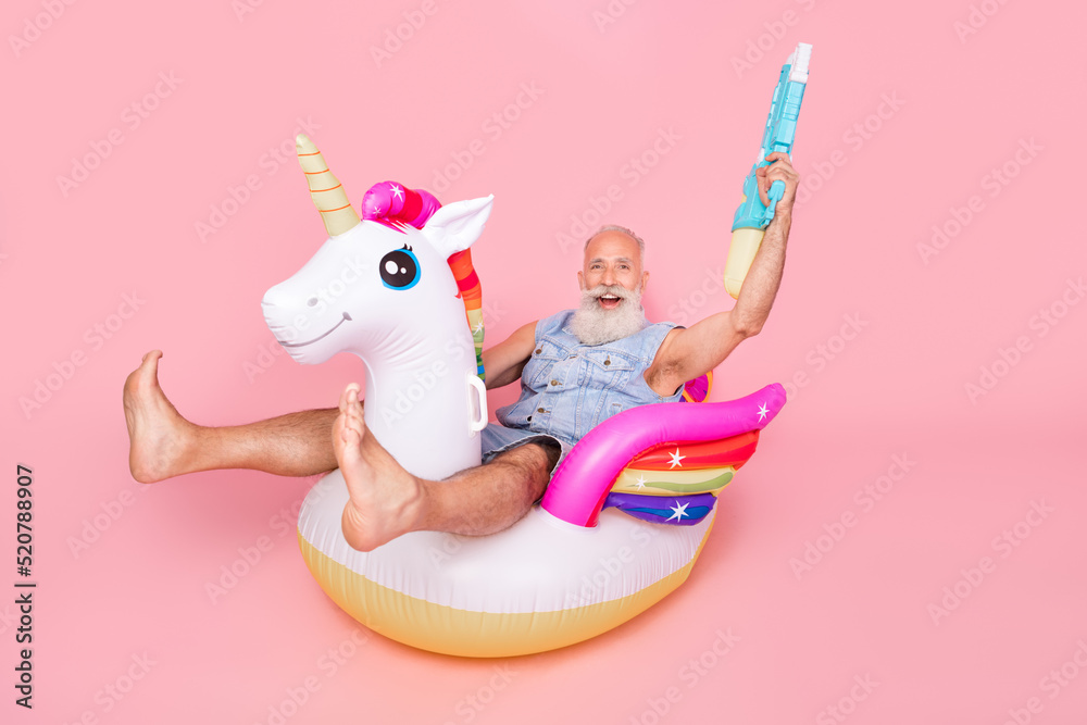 Full length photo of pretty excited retired guy wear jeans waistcoat rising water weapon sitting unicorn circle isolated pink color background