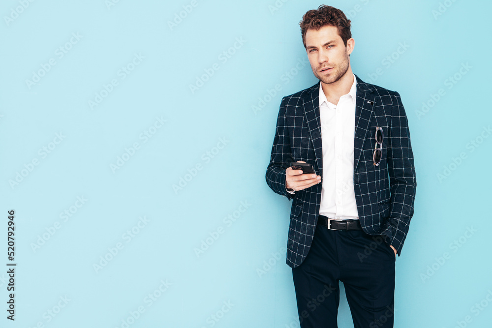 Portrait of handsome confident stylish hipster lambersexual model. Sexy modern man dressed in black elegant suit. Fashion male posing in studio near blue wall. Holding mobile phone smartphone Portrait of handsome confident stylish hipster lambersexual model. Sexy modern man dressed in black elegant suit. Fashion male posing in studio near blue wall. Holding mobile phone smartphone
