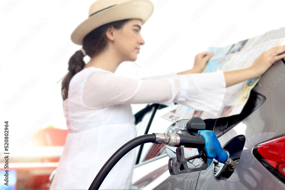 Obraz premium Blue fuel petrol pump nozzle against refueling car with petrol with beautiful female traveler holding touristic map and waiting as blurred background at gas station, customer woman self-refueling car.