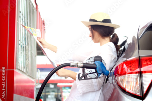 Blue fuel petrol pump nozzle against refueling auto car with petrol, beautiful female traveler holding touristic map, waiting as blurred background at gas station, customer woman self-refueling car.