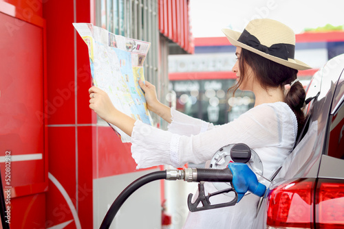 Blue fuel petrol pump nozzle against refueling auto car with petrol, beautiful female traveler holding touristic map, waiting as blurred background at gas station, customer woman self-refueling car.