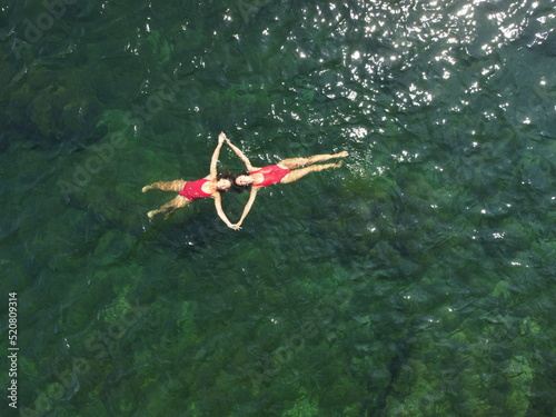 Woman floating sea. Aerial top view of two pretty woman in red swimsuit floating on water surface in crystal clear turquoise sea. Vacation at Paradise. Ocean relax, travel and vacation. Slow motion