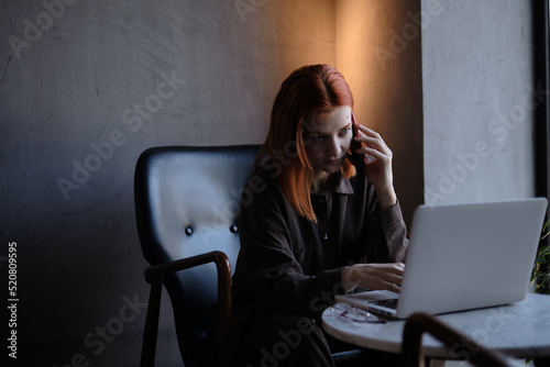 a red-haired freelance woman with glasses works remotely on a laptop in a cafe.x