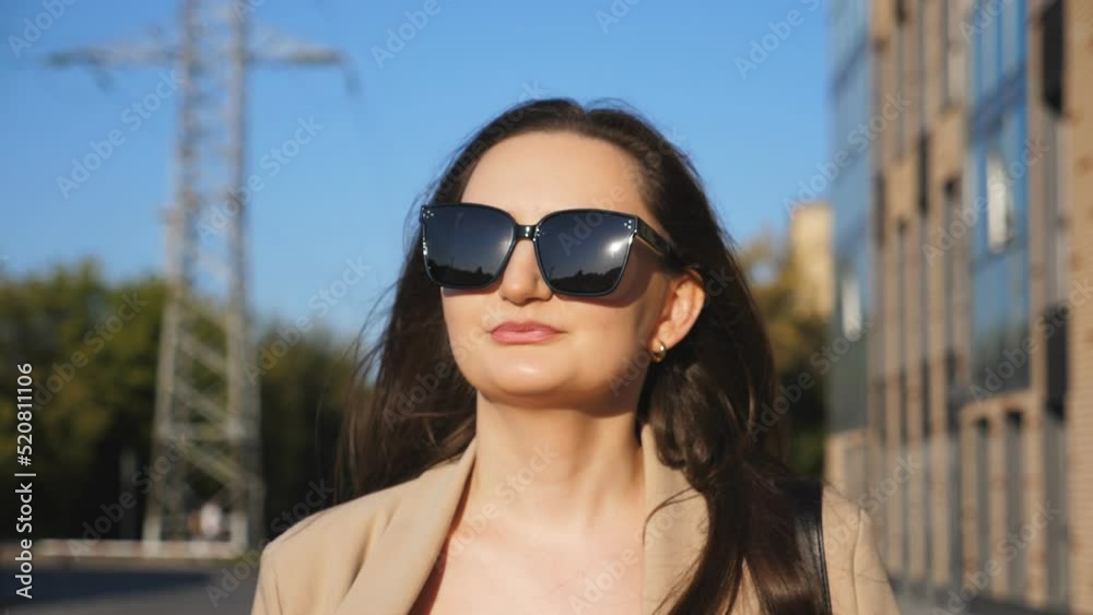 Portrait of beautiful lady in sunglasses walking at sidewalk. Attractive brunette girl looks into camera going along city street. Businesswoman commuting to work. Blurred urban background. Close up Portrait of beautiful lady in sunglasses walking at sidewalk. Attractive brunette girl looks into camera going along city street. Businesswoman commuting to work. Blurred urban background. Close up