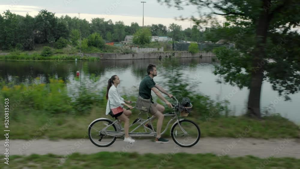 A young married couple, a man and a woman, ride together on a two-seater urban tandem bike along the road along the river bank A young married couple, a man and a woman, ride together on a two-seater urban tandem bike along the road along the river bank