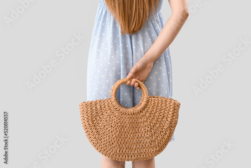 Young woman holding rattan bag behind her back on light background