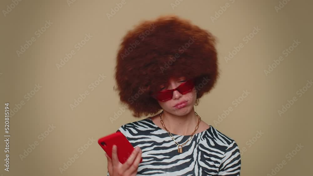 Sad young woman with brown lush wig use smartphone typing browsing, loses becoming surprised sudden lottery results, bad fortune, loss. Adult stylish girl isolated on beige studio background indoors Sad young woman with brown lush wig use smartphone typing browsing, loses becoming surprised sudden lottery results, bad fortune, loss. Adult stylish girl isolated on beige studio background indoors