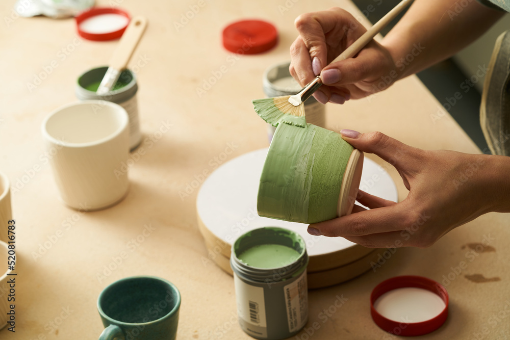 Close-up of girl painting clay mug with glaze. Woman coloring pottery in workshop with a paintbrush. Painter in green apron glazing clay pot - obrazy, fototapety, plakaty 
