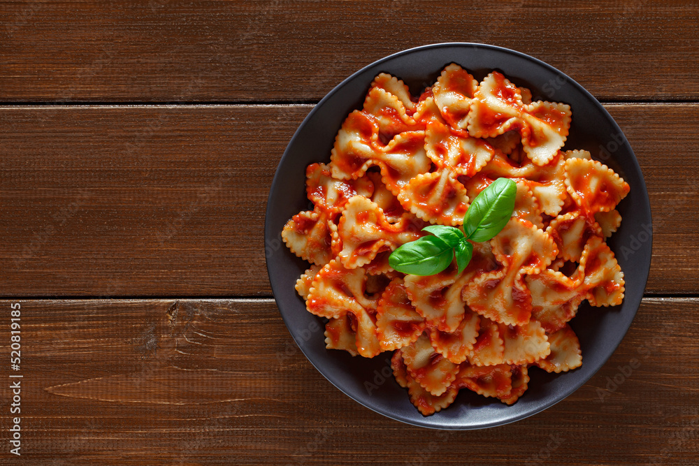 Farfalle, cooked pasta with tomato sauce and basil green leaf, in gray plate, on wooden brown plank tabletop background, top view, space to copy text.. - obrazy, fototapety, plakaty 