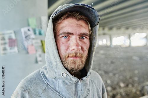 Close-up of bearded homeless man with dirty face in torn clothes looking at camera standing outdoors