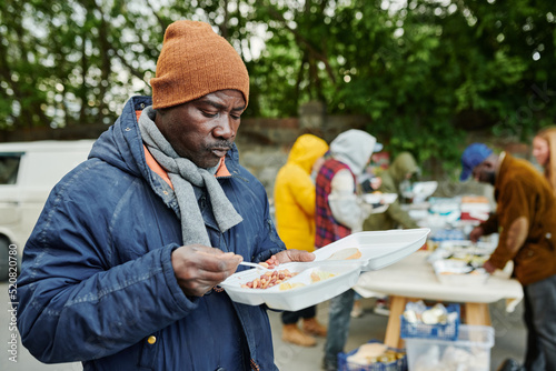 African poor man in warm clothing eating food giving by volunteers outdoors for homeless people photo