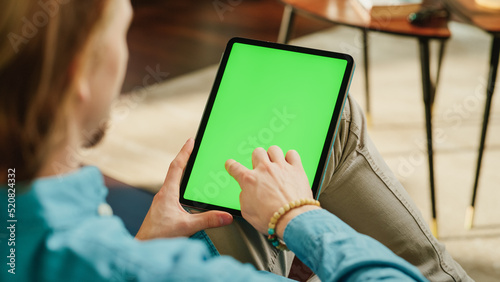 Young Man Scrolling Content on Tablet Computer with Green Screen Mock Up Display. Male Relaxing at Home, Reading Social Media Posts on Mobile Device. Close Up Over the Shoulder Photo.