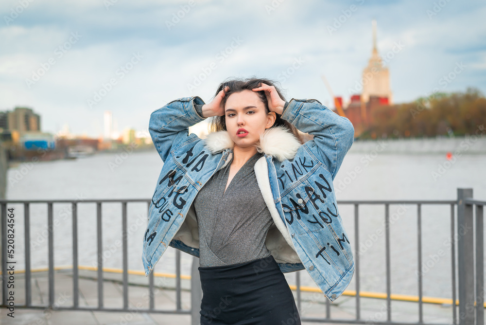 Fototapeta premium A beautiful girl, dressed in a jeans jacket, walks along the pier on the banks of the river.
