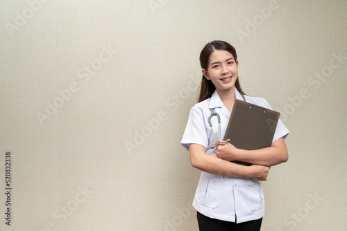 A long-haired brown-haired Asian female doctor in a white short-sleeved medical uniform smiling brightly, is standing with a paper board isolated against the background.