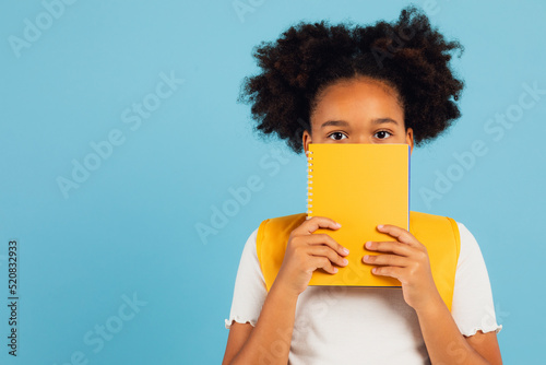 Funny african american schoolgirl covering her face with yellow book on blue background, back to school concept.