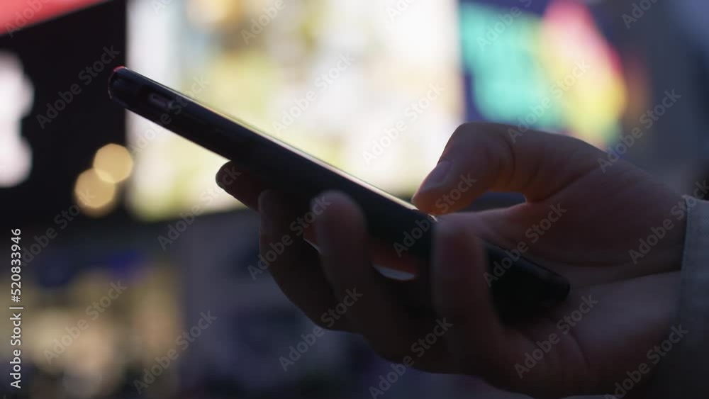 Close up of teenagers hand in the city using a cell phone with background of bright lights and screens Close up of teenagers hand in the city using a cell phone with background of bright lights and screens