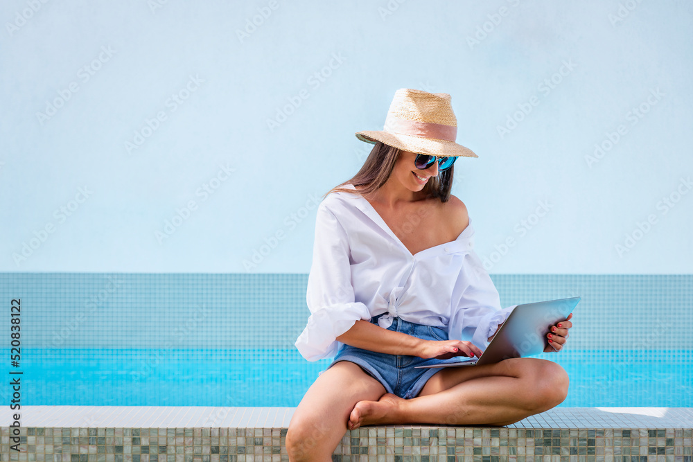 Happy woman using a laptop while sitting at swimming pool and browsing on the internet