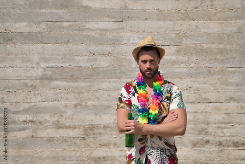 Young and handsome man, blue eyes, with beard, hat, Hawaiian shirt and flower necklace with arms crossed and a bottle of beer in his hand on a gray background. Concept holidays, party, trips, drink.
