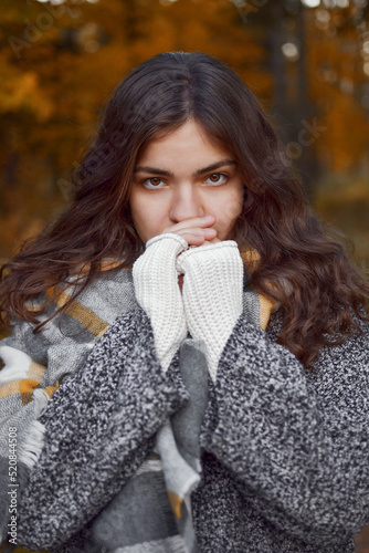 Portrait of woman with curly brown hair looking straight at camera covering lower part of face with hands to keep warm, checkered scarf.