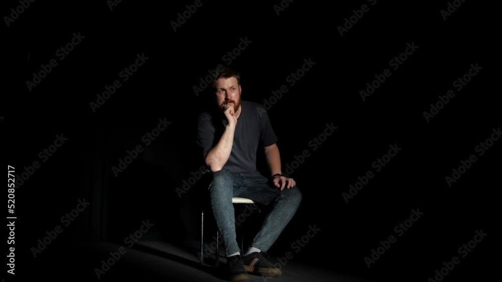 A bearded man sits thoughtfully on a chair in a dark room illuminated by a beam of light from an open door or window. The guy podsve Dramatic scene, loneliness, hope. A bearded man sits thoughtfully on a chair in a dark room illuminated by a beam of light from an open door or window. The guy podsve Dramatic scene, loneliness, hope.