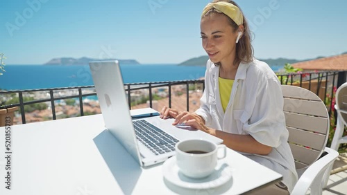 Young elegant woman with wireless earphones looking at laptop with smile while sitting at balcony with seaview on resort. girl freelancer works outdoors during travel