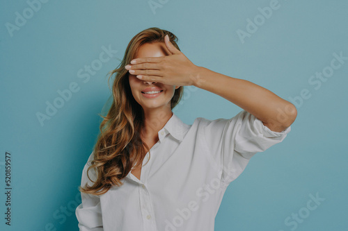 Portrait of cheerful young woman broadly smiling while covering her eyes with hand