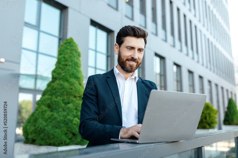 Smiling confident millennial attractive european businessman with beard in suit chatting on laptop