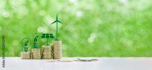 Renewable or clean energy generation prices and costs, financial concept : Green eco-friendly symbols atop coin stacks e.g. energy efficient light bulb, a battery, a solar cell panel,  a wind turbine.