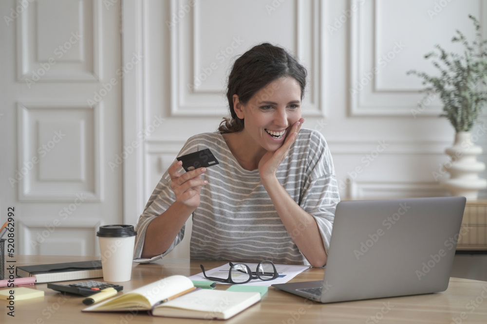 Exited Italian entrepreneur lady joyfully looking at computer screen holding credit card Exited Italian entrepreneur lady joyfully looking at computer screen holding credit card
