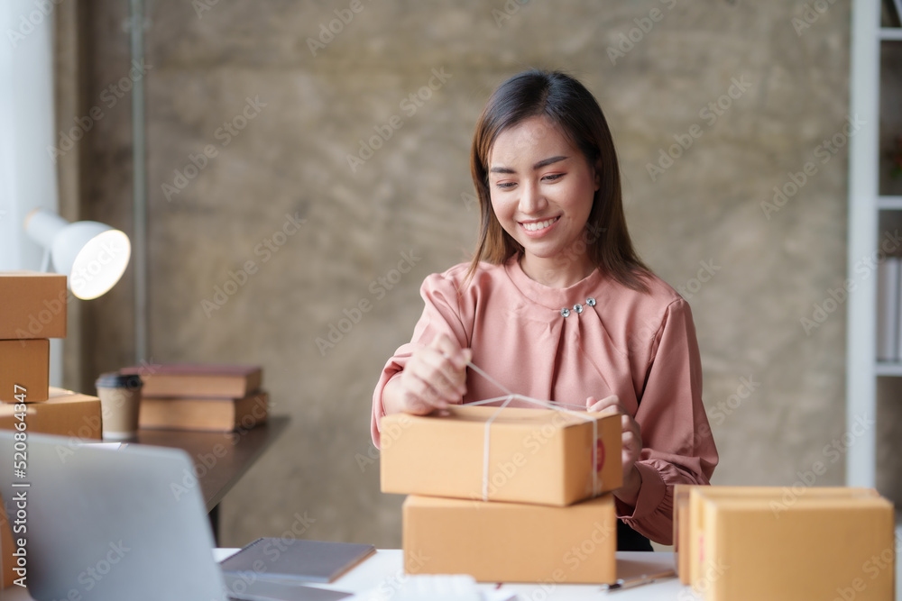 Smiling Asian online business owner prepares parcels and checks online order for delivery to customer on his laptop computer. Online selling concept. Smiling Asian online business owner prepares parcels and checks online order for delivery to customer on his laptop computer. Online selling concept.