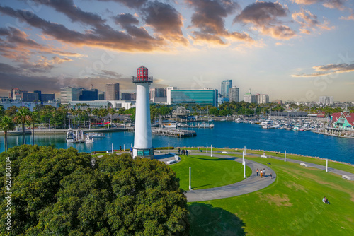 a gorgeous summer landscape in the park with a tall white and red lighthouse surrounded by lush green palm trees and grass with skyscrapers in the city skyline and blue ocean water at sunset