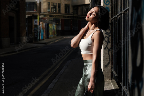 A young asian woman is standing in a street of London and enjoying morning sun.