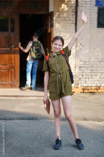 a teenage girl  being a student  stands next to the entrance to the school building  waving a greeting