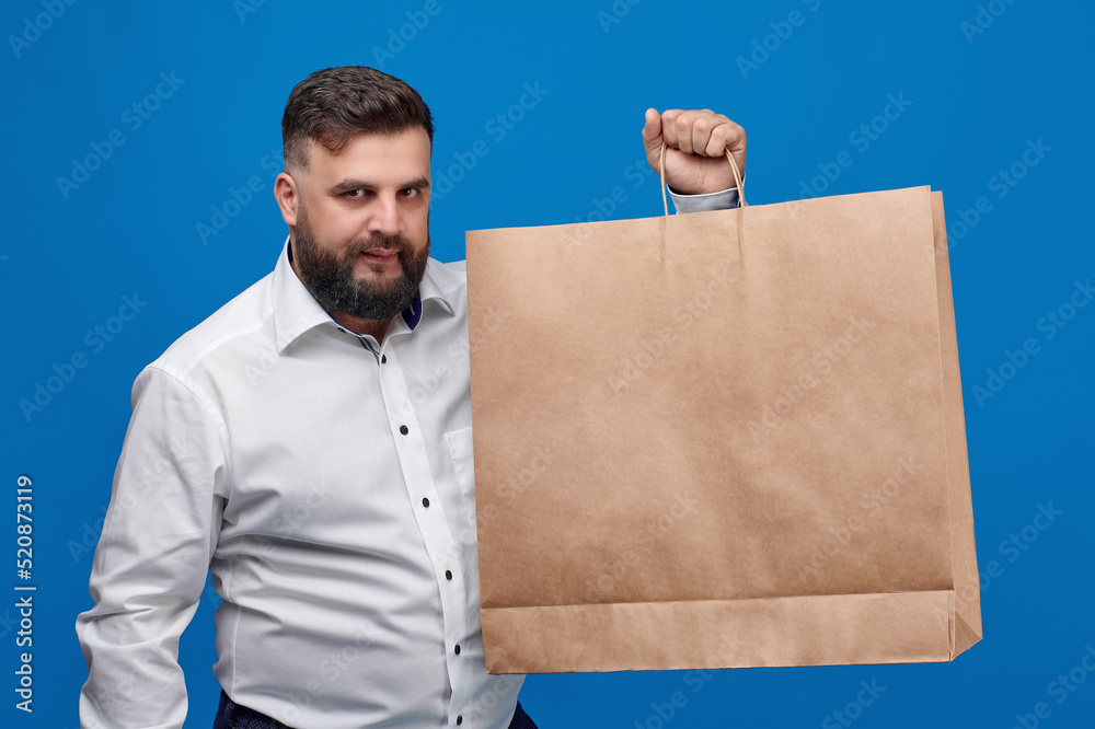 A Caucasian-looking man holds a large brown paper bag. A man in a white shirt with shopping bags on a blue background. Space for text on the shopping banner ad. Banner for discounts. A Caucasian-looking man holds a large brown paper bag. A man in a white shirt with shopping bags on a blue background. Space for text on the shopping banner ad. Banner for discounts.