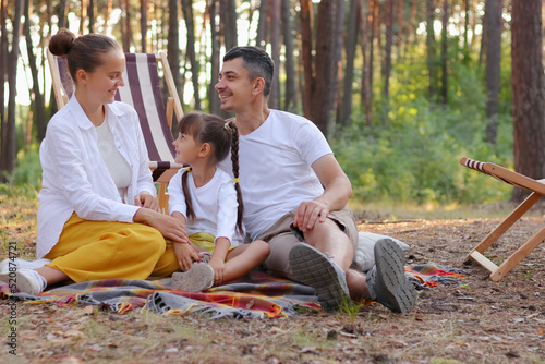 Outdoor portrait of smiling delighted family spending time together in the forest, people wearing casual clothing, sitting on blanket and talking with each other with pleasure. © sementsova321